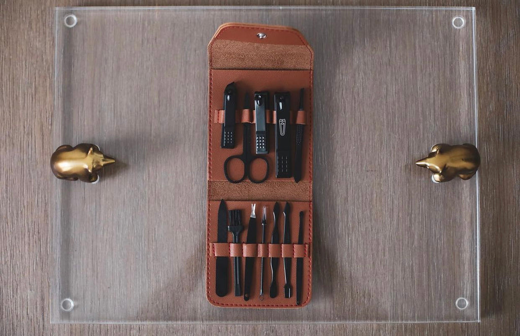 A collection of nail grooming tools including nail clippers, file, scissors, tweezers, cuticle pusher, and other items, neatly arranged in an open brown case. 